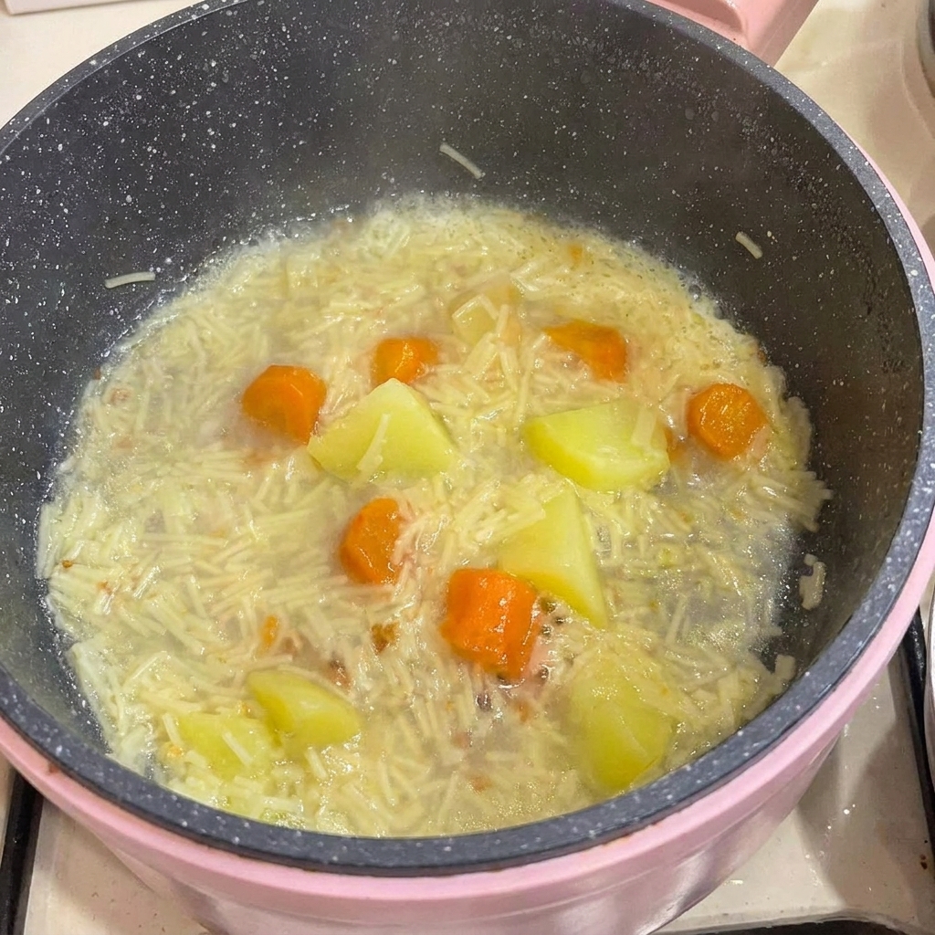 Deboned chicken meat being added into a pot filled with cooked baby noodles, potatoes, and carrots in broth.
