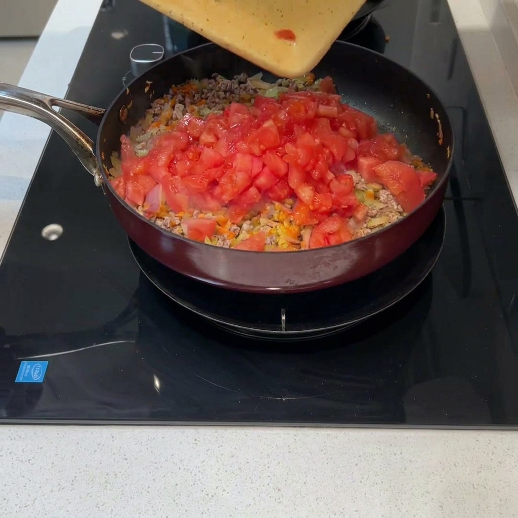 Freshly diced tomatoes being poured from a cutting board into a pan with meat and vegetables.