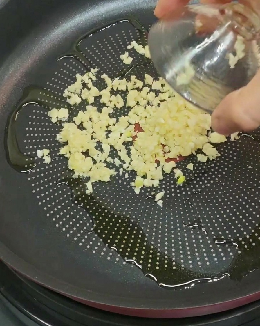Finely minced garlic pieces being poured from a small glass bowl into a frying pan containing olive oil.