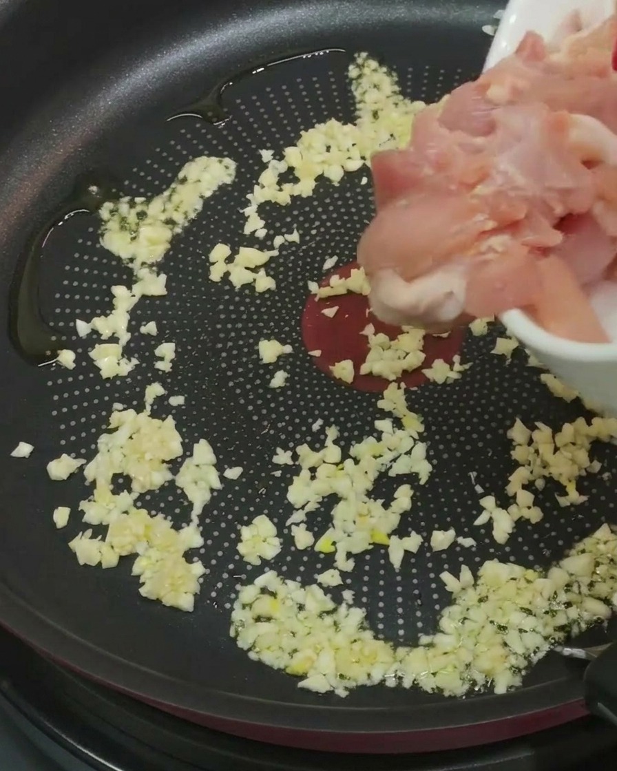 A handful of raw, marinated chicken pieces being added to a frying pan already containing minced garlic and oil.