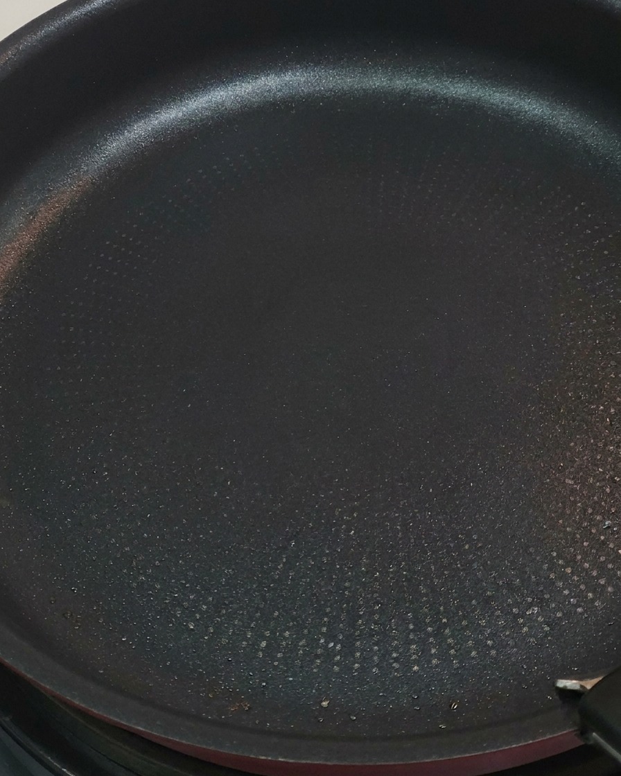 Red tongs flipping pieces of chicken in a frying pan, showing some surfaces turned to a golden brown color.