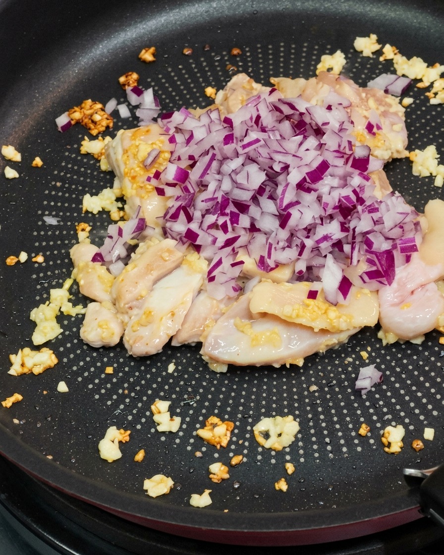 Diced red onions being poured into a pan with sautéed chicken and minced garlic.