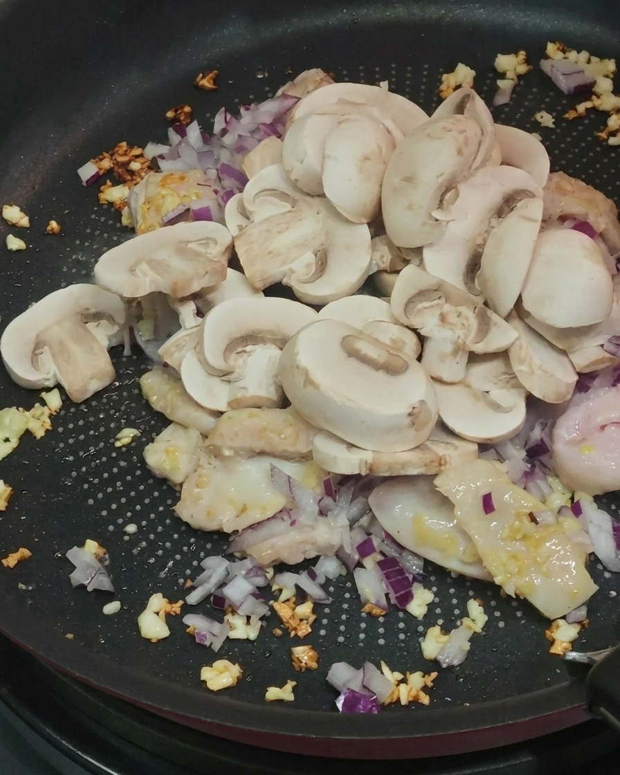 Freshly sliced white button mushrooms being added to a pan containing cooked chicken, garlic, and red onions.