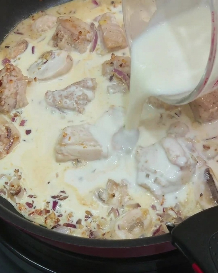 Pouring milk from a glass measuring cup into a pan filled with cooked chicken pieces, sliced mushrooms, onions, and cream.