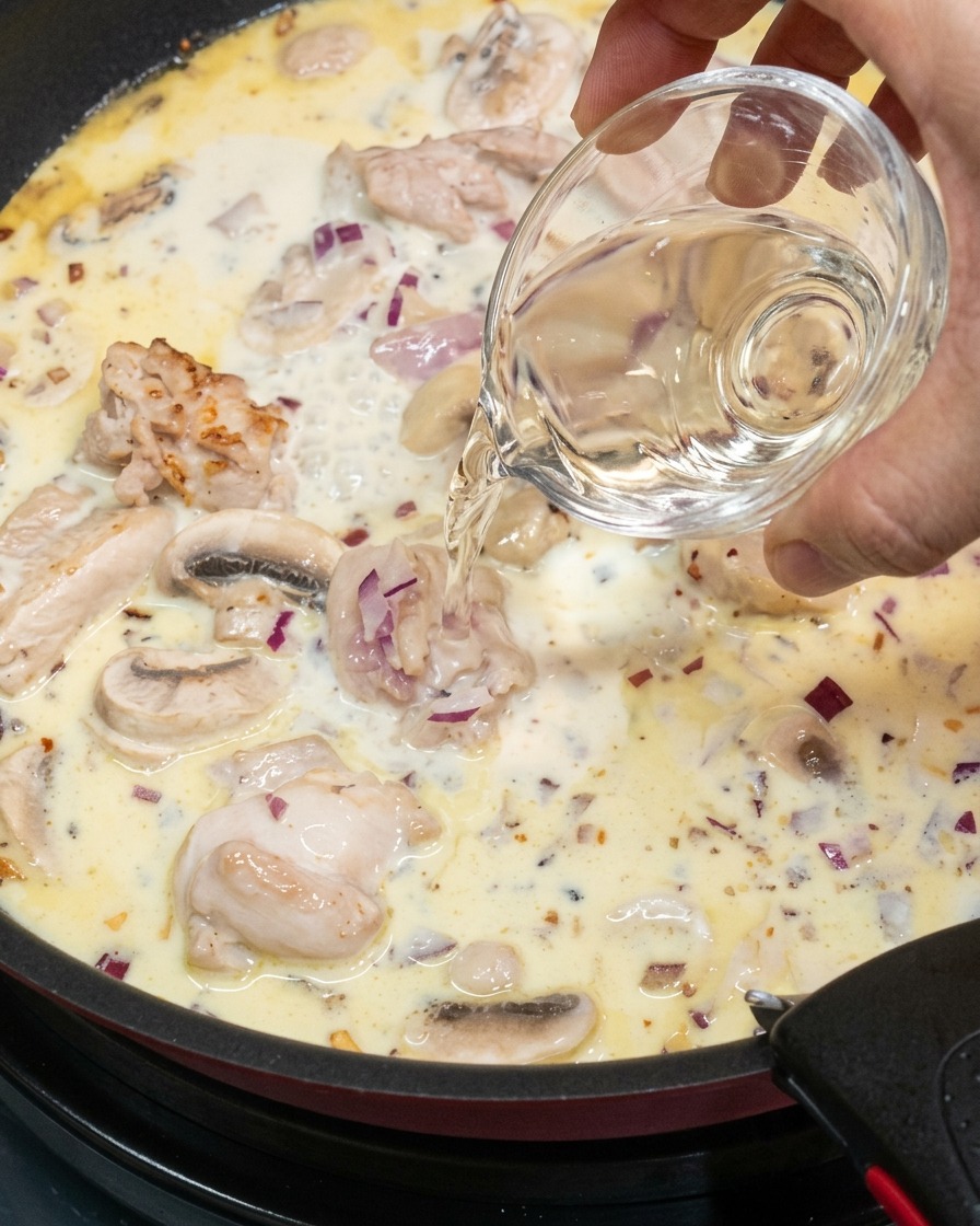 A hand pouring white wine from a small glass bowl into a creamy, simmering sauce with chicken and mushrooms.