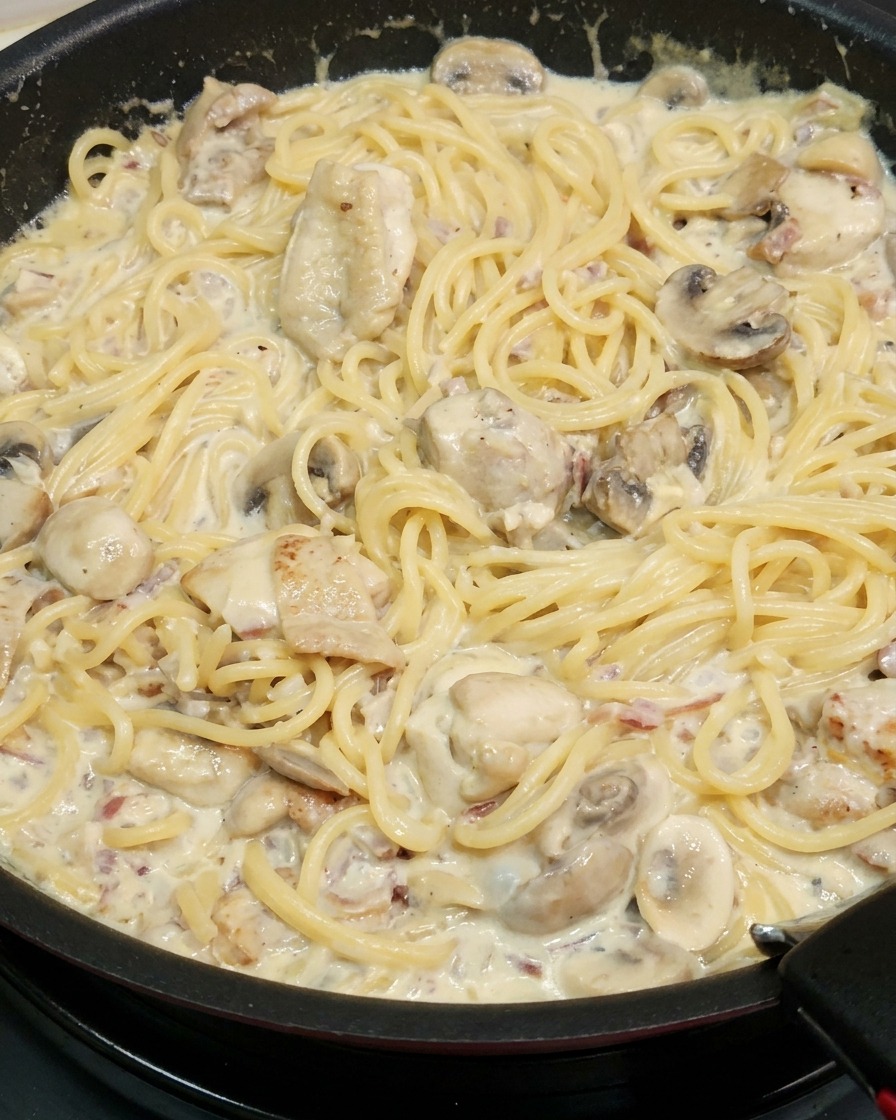 Cooked spaghetti noodles being tossed in a pan with a rich, thickened creamy sauce, chunks of chicken, and sliced mushrooms.