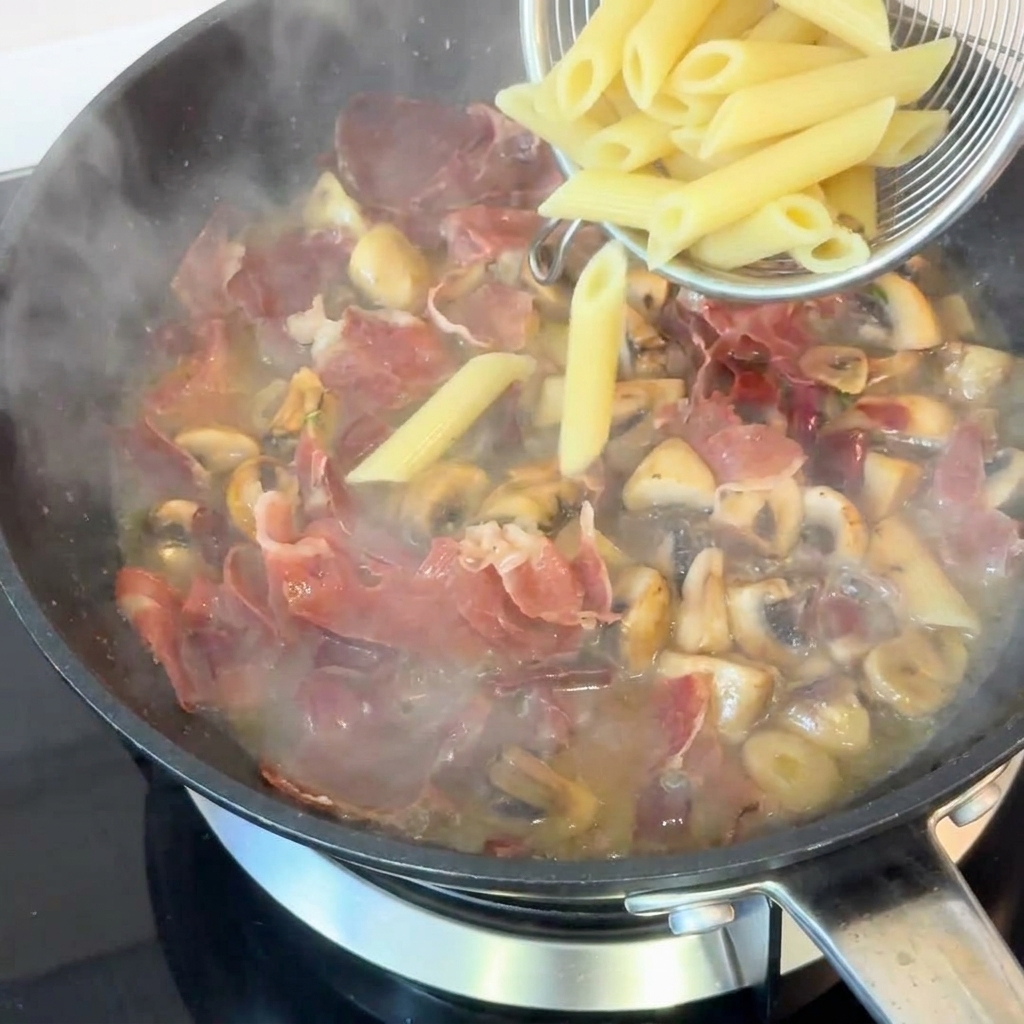 Cooked penne pasta being scooped with a metal spider strainer and dropped into a pan full of savory ham, mushrooms, and rich pan sauce.