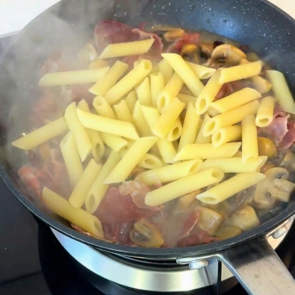 Cooked penne pasta being tossed in a pan with sautéed mushrooms and ham, seasoned with black pepper.