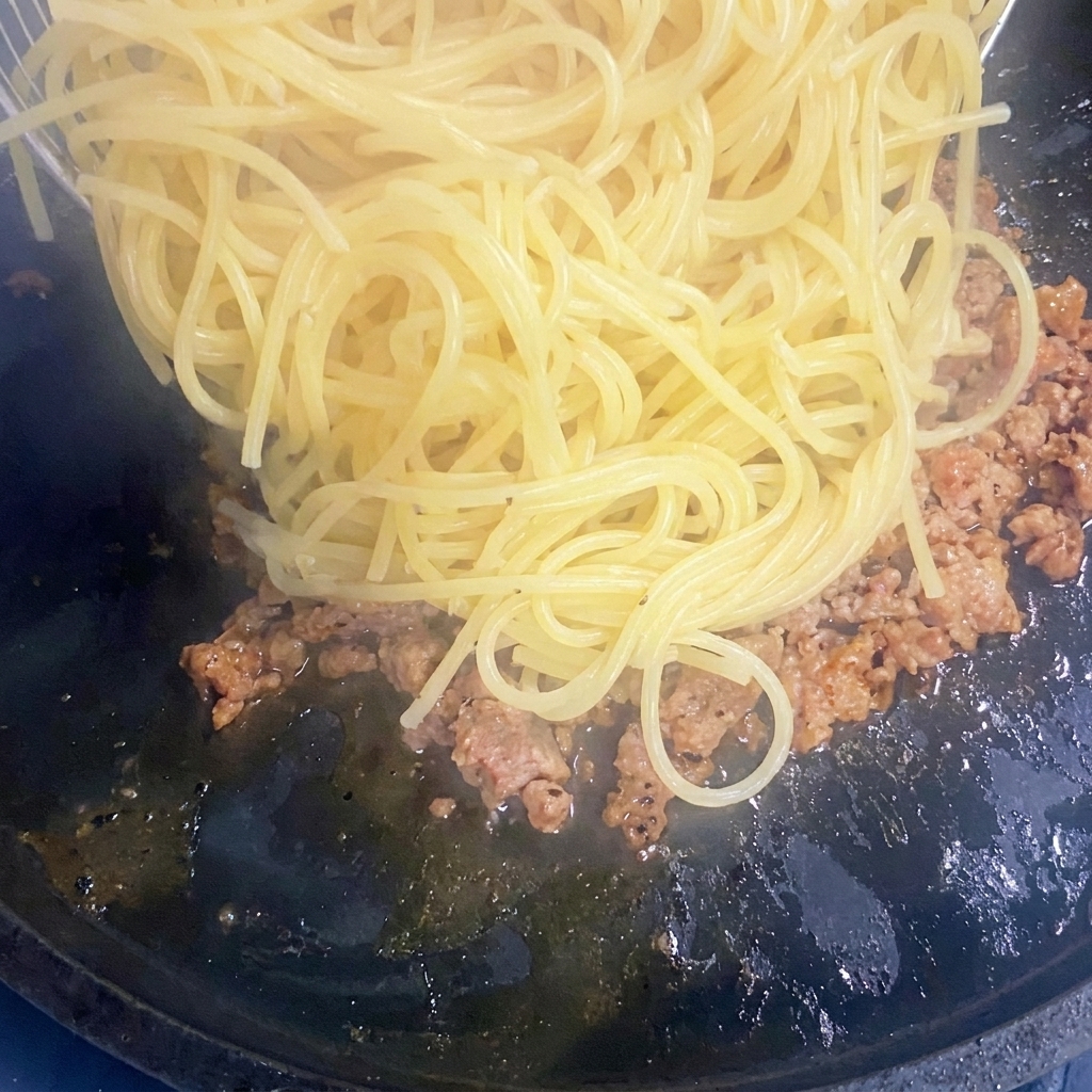 Cooked spaghetti noodles being added to a pan of browned ground meat.