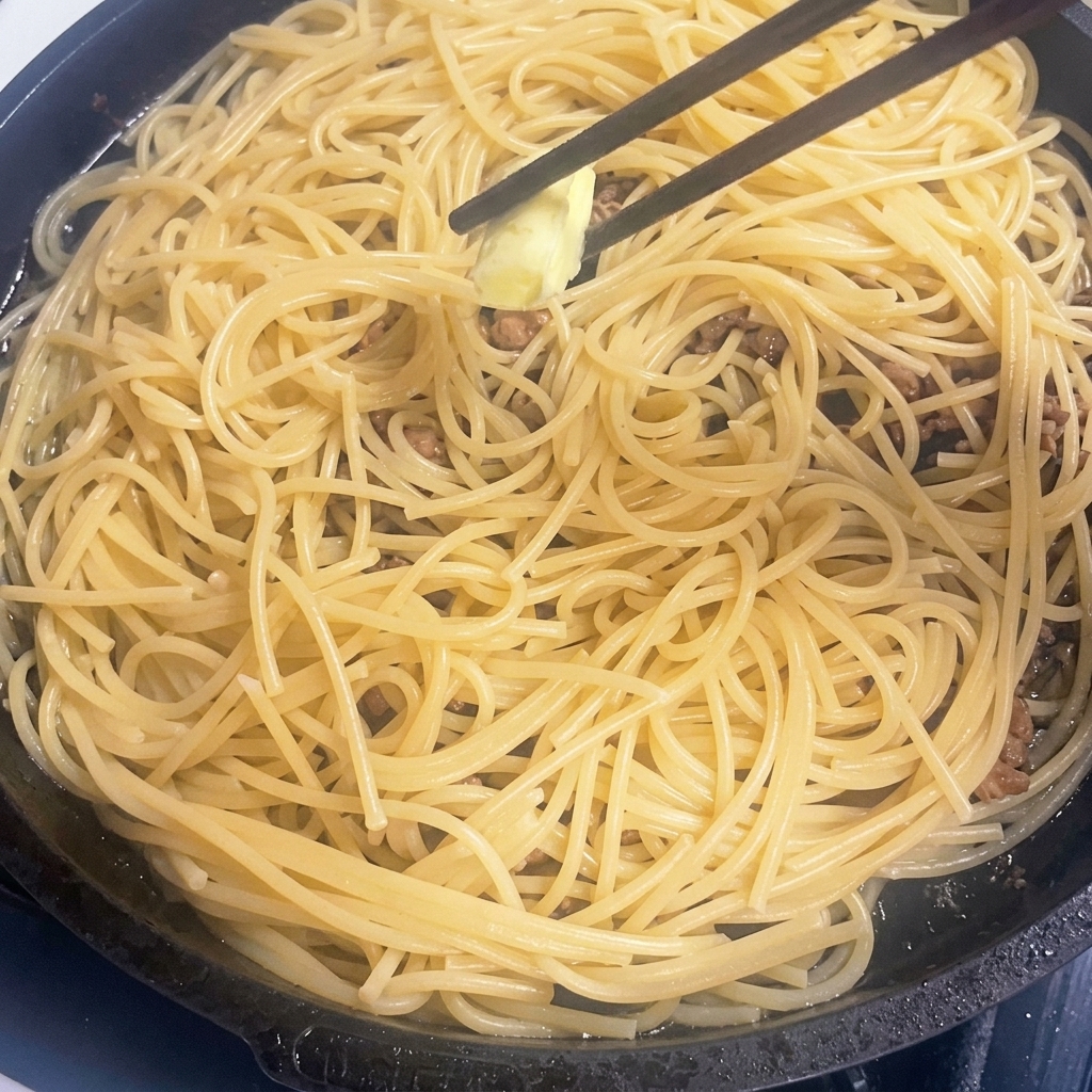 A small piece of butter melting into a pan of hot pasta and meat.