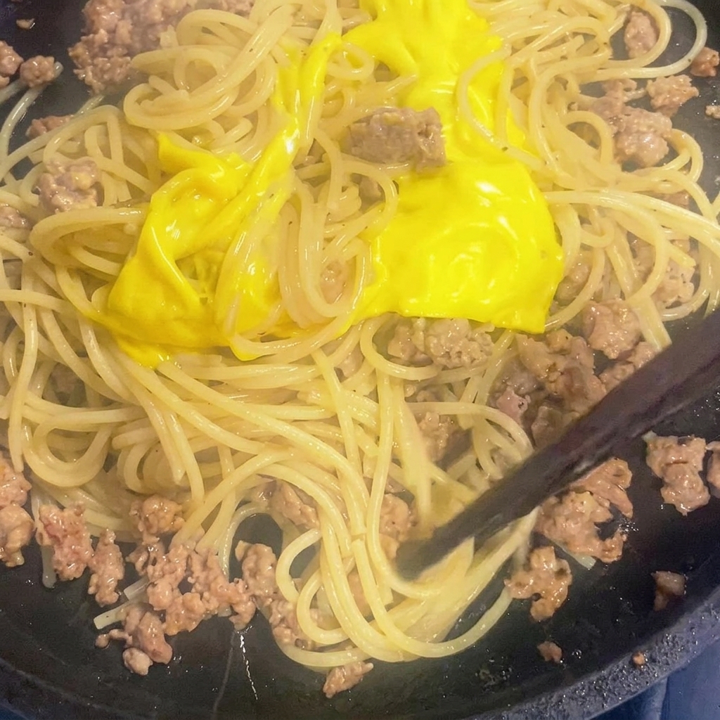 Melted cheese being stirred into spaghetti and meat sauce in a skillet.
