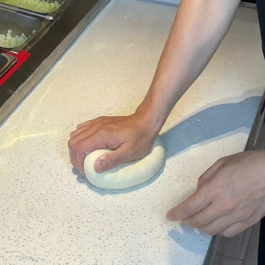 Hands pressing down on a round ball of pizza dough on a floured white countertop.