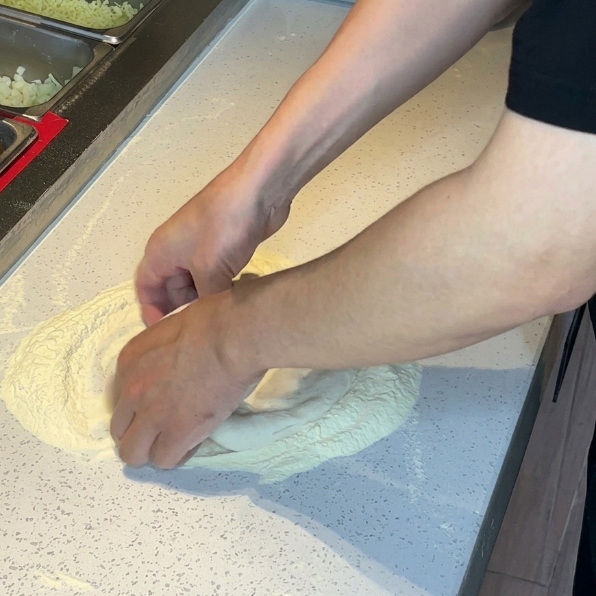 Hands pressing and stretching pizza dough in a pile of flour on a white surface.