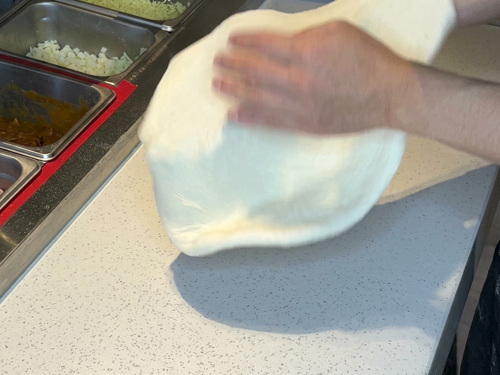 Two hands lifting and tossing stretched pizza dough in the air to remove excess flour.