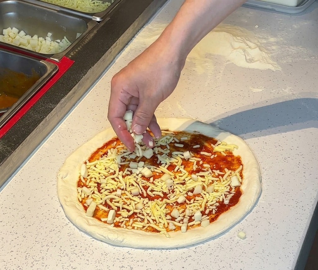 A hand sprinkling shredded white cheese over a pizza crust spread with tomato sauce.