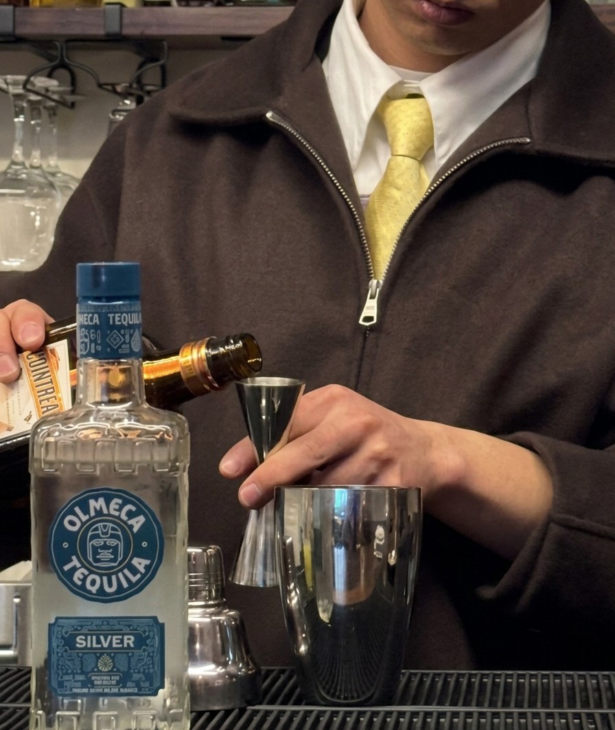 Triple sec being poured from a bottle into a jigger positioned over a shaker tin.