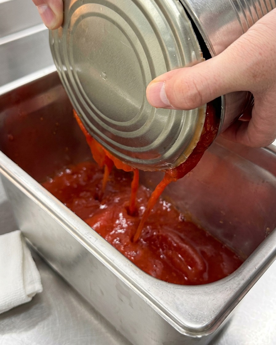Peeled canned tomatoes being poured into a clean stainless steel catering pan.