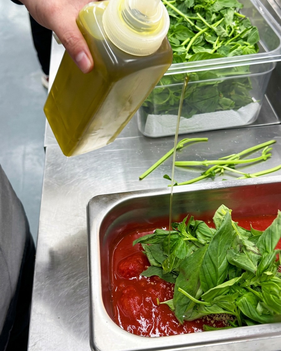 Olive oil being drizzled from a bottle into a container of mixed tomato sauce ingredients.