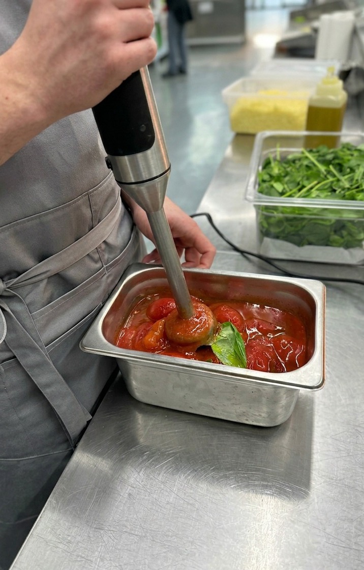 A close-up of an immersion blender processing whole peeled tomatoes and basil leaves into a smooth, vibrant red pizza sauce.