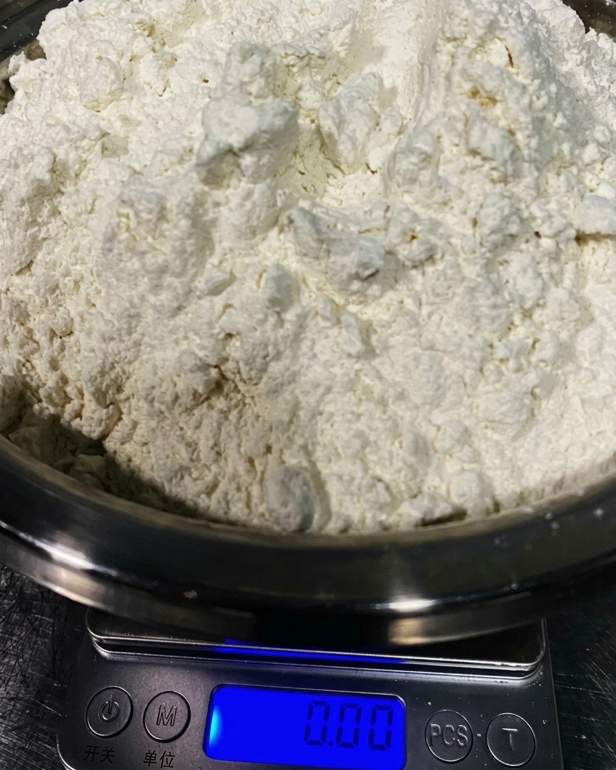 A large mound of fine white 00 flour sitting in a stainless steel bowl on a kitchen scale.