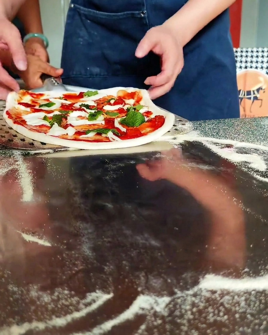 Hands positioning a raw, topped pizza onto a perforated metal peel on a flour-dusted countertop.