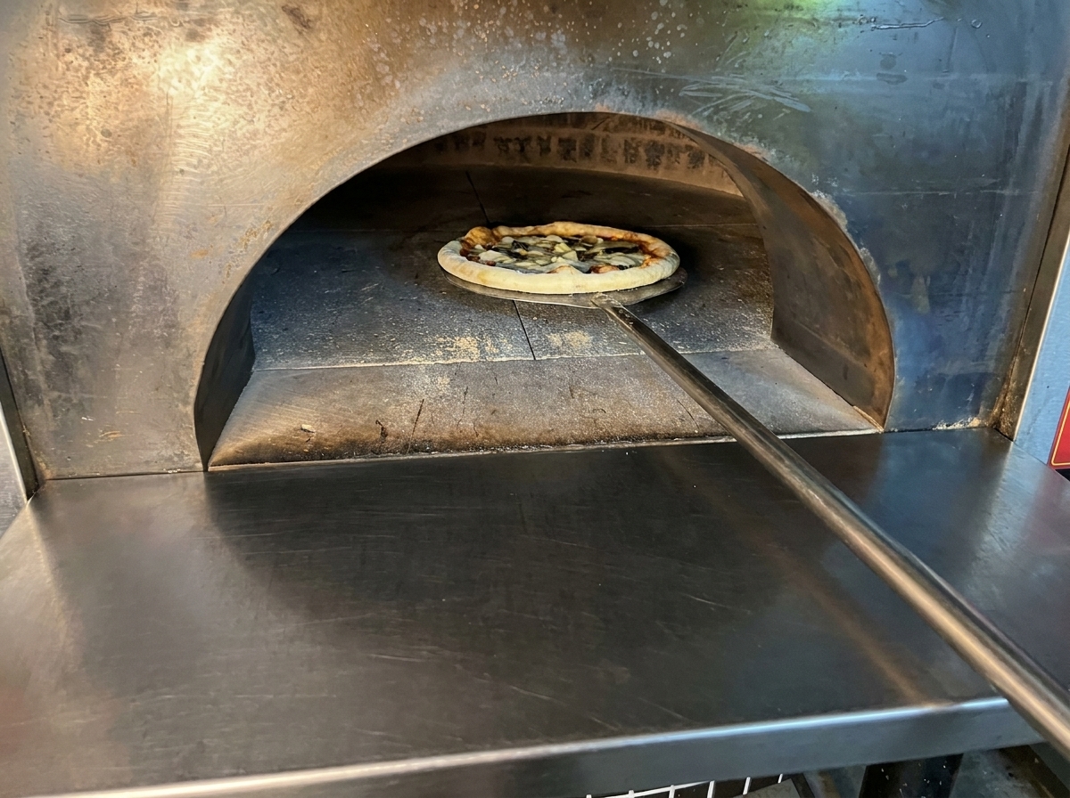 Assembled raw pizza being placed into a hot wood-fired oven using a long metal peel.