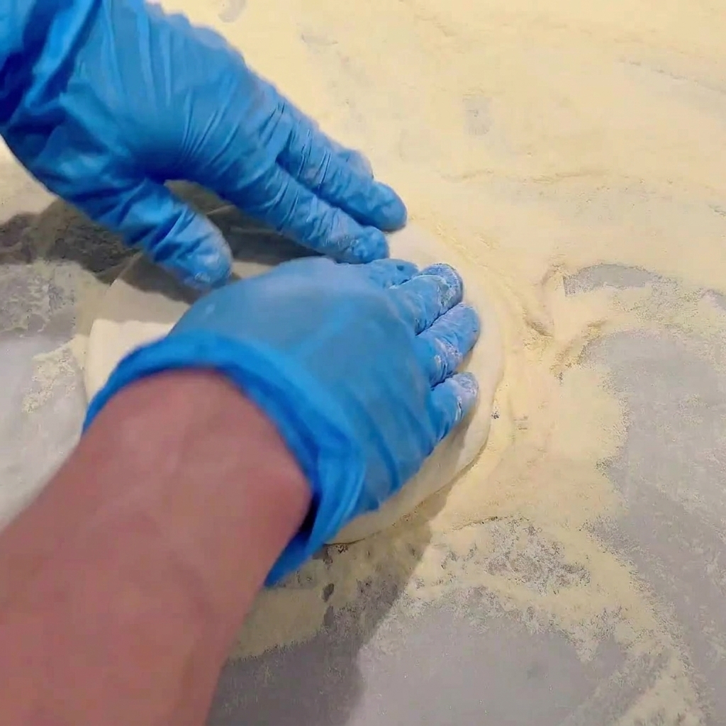 Hands wearing blue gloves pressing and stretching raw pizza dough on a flour-dusted countertop.