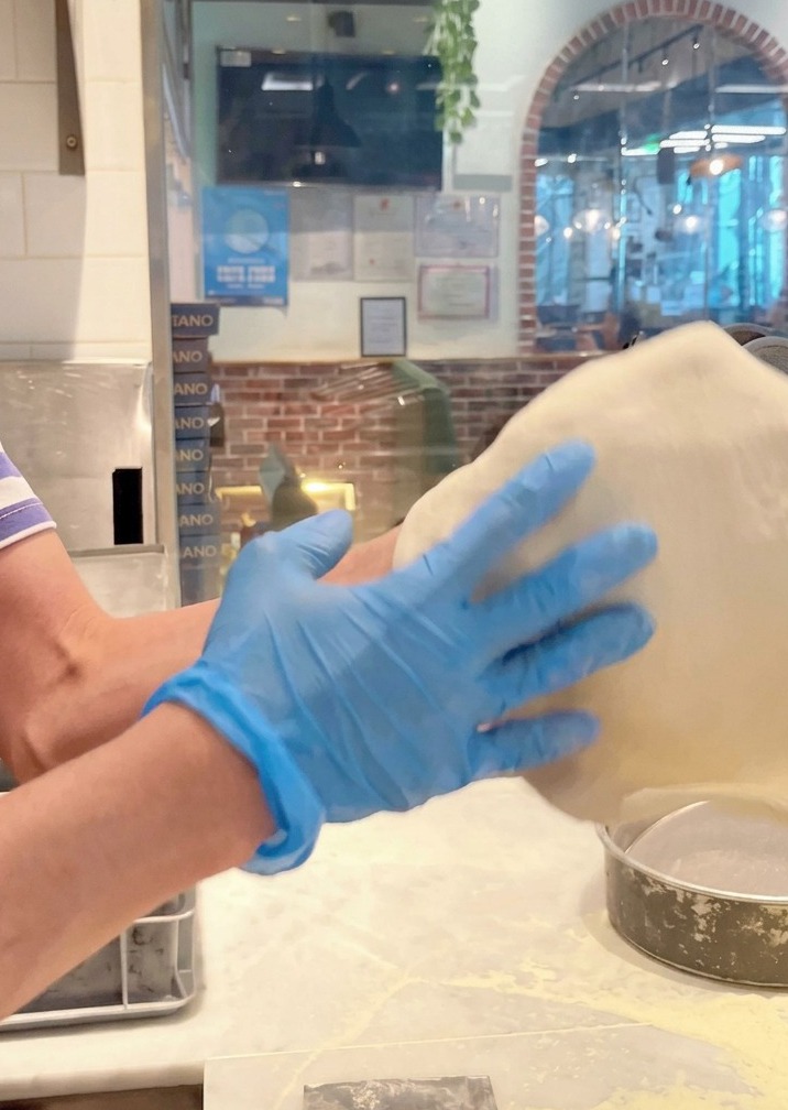 Pizza dough being tossed and stretched in the air by hands wearing blue gloves.