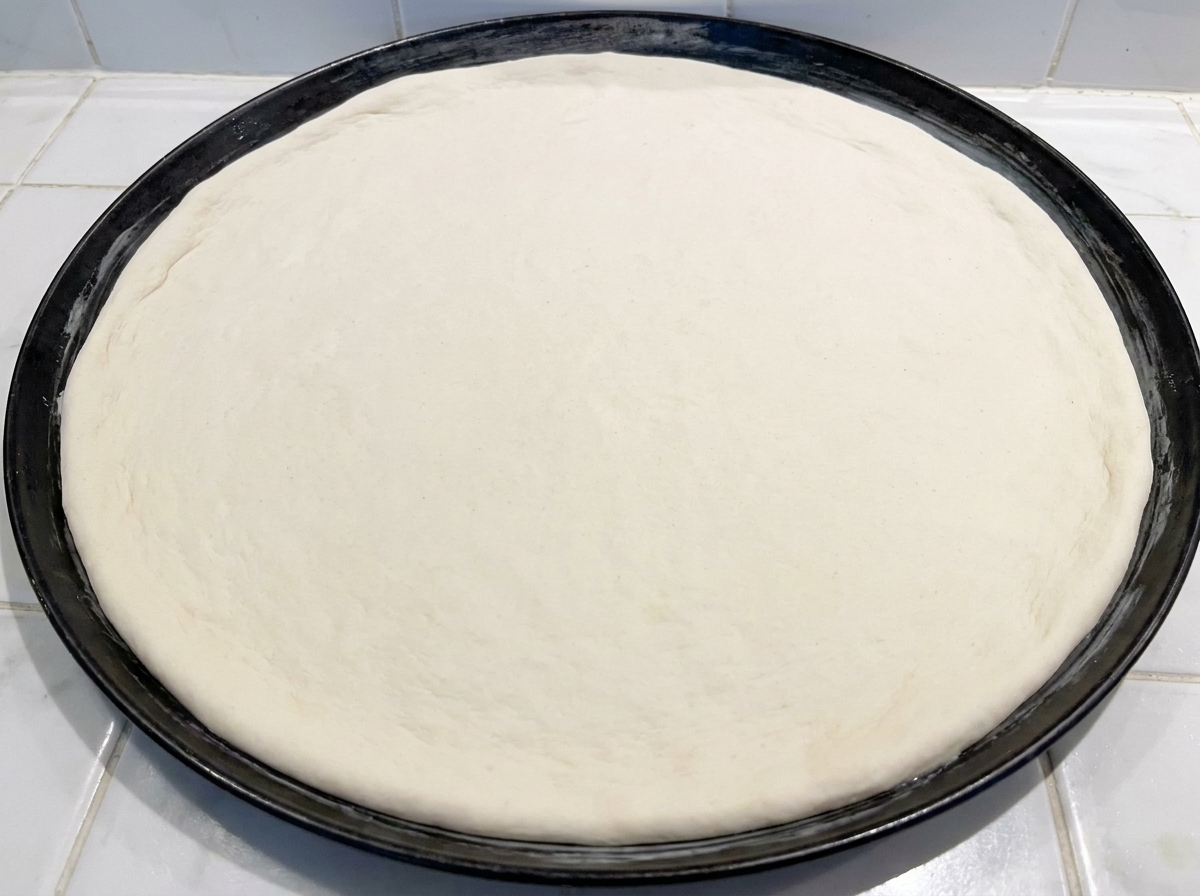A round of raw pizza dough being pressed into a black metal baking pan.