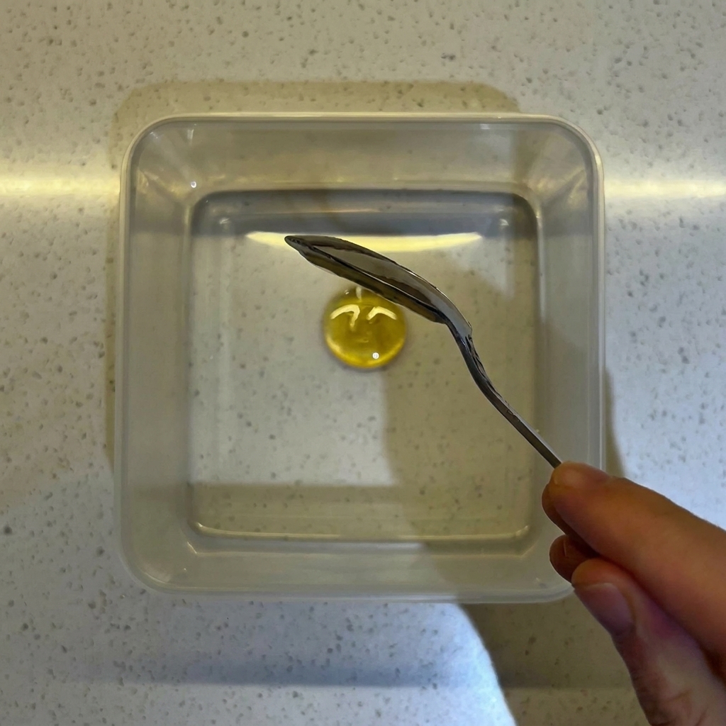 A close-up of a spoon mixing honey and yeast into water in a clear square container.