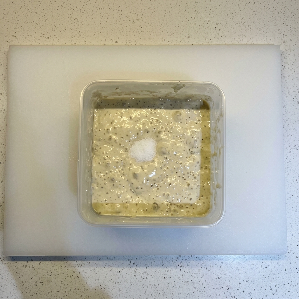 Salt and a heap of flour sitting on top of a bubbly, fermented dough mixture in a square container.