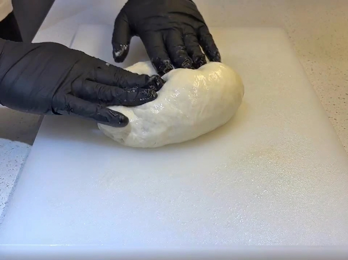 Black-gloved hands folding a soft, oiled dough ball on a white cutting board.