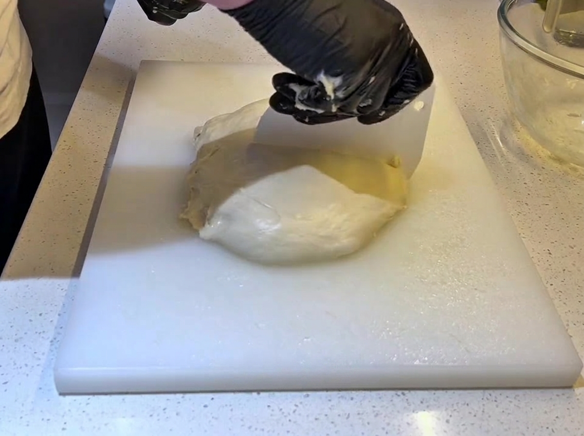 Black-gloved hands using a white plastic dough scraper to cut a large piece of dough in half.