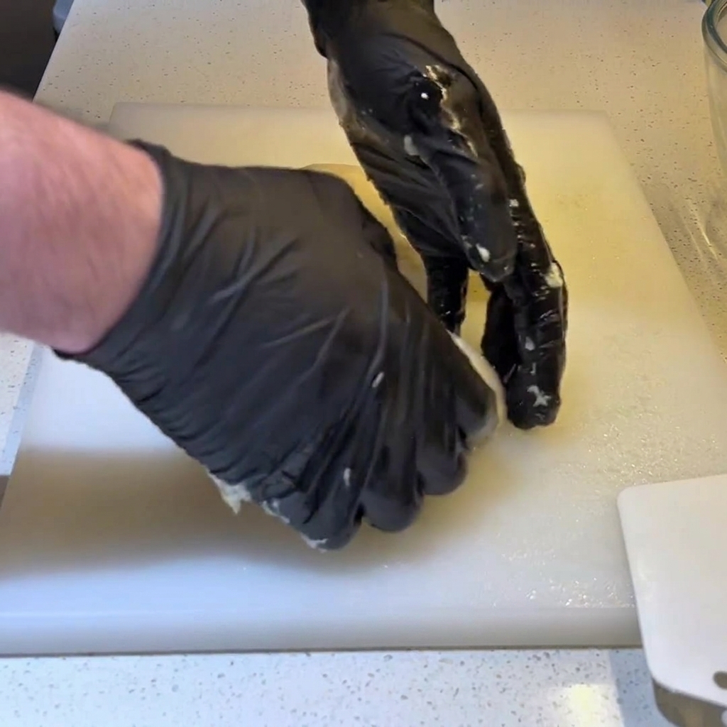 Gloved hands tucking the edges of a dough portion underneath to form a tight, round ball.