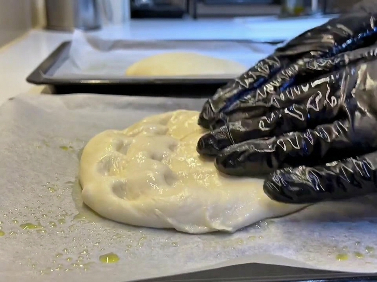 Gloved hands gently pressing and stretching a piece of soft dough into a rectangle on oiled parchment paper.
