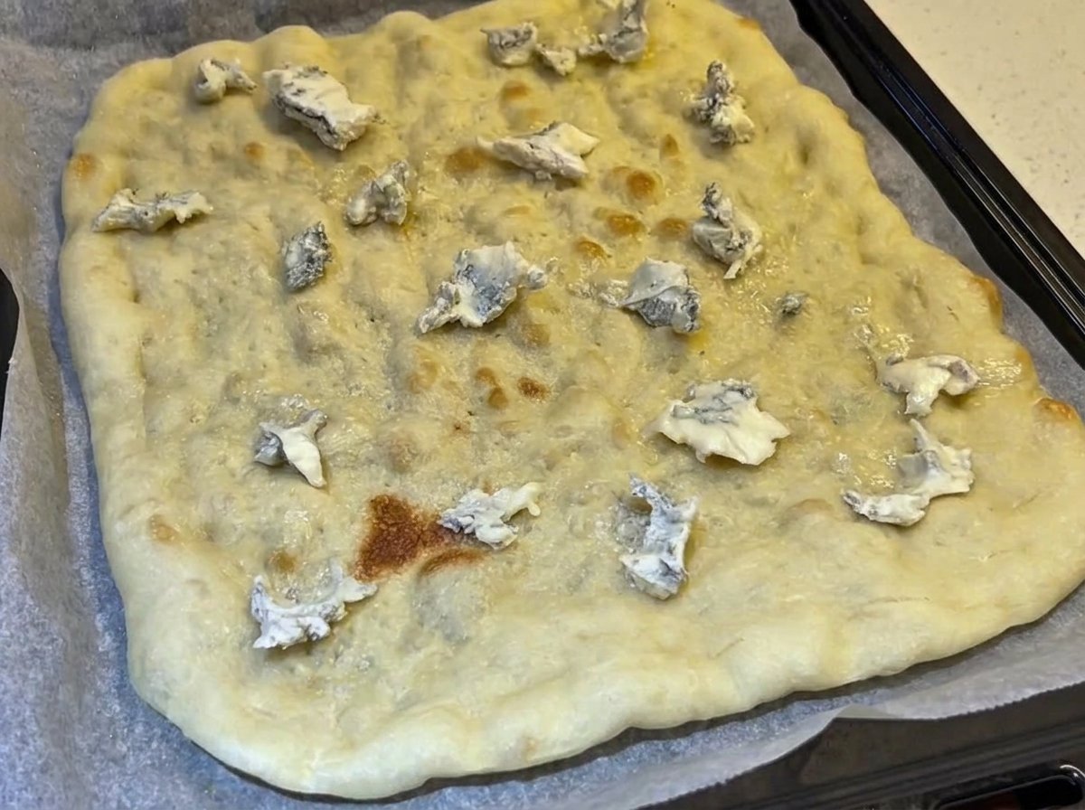 A pre-baked rectangular flatbread crust topped with scattered crumbles of blue cheese on a parchment-lined baking tray.