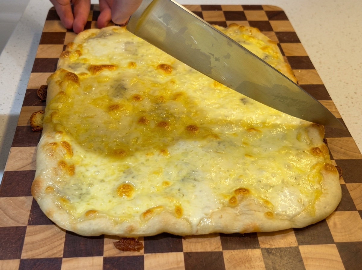 A large chefs knife slicing through a freshly baked, bubbling four-cheese rectangular pizza on a checkered wooden cutting board.