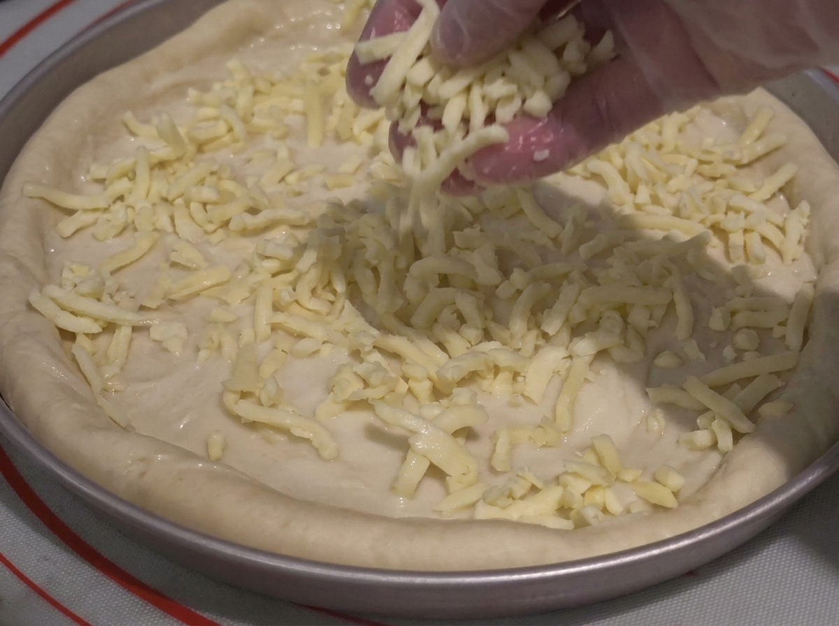 A gloved hand sprinkling shredded cheese generously over a sauce-covered pizza dough in a pan.