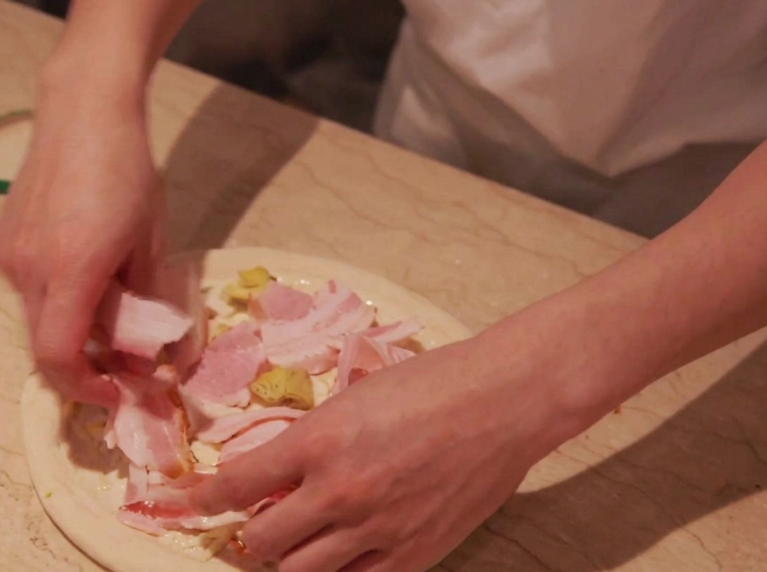 Slices of pork being carefully layered over the potato topping on the pizza dough.