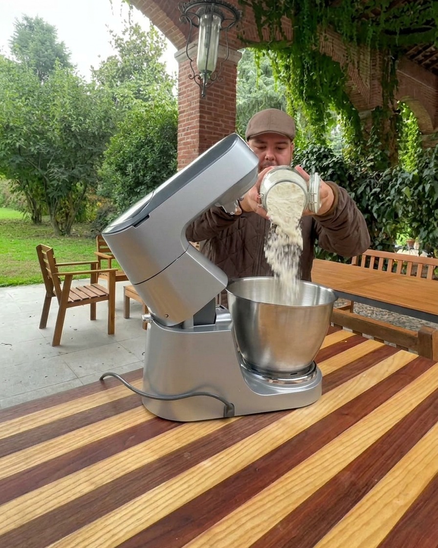 Flour being poured into a large stainless steel stand mixer bowl.