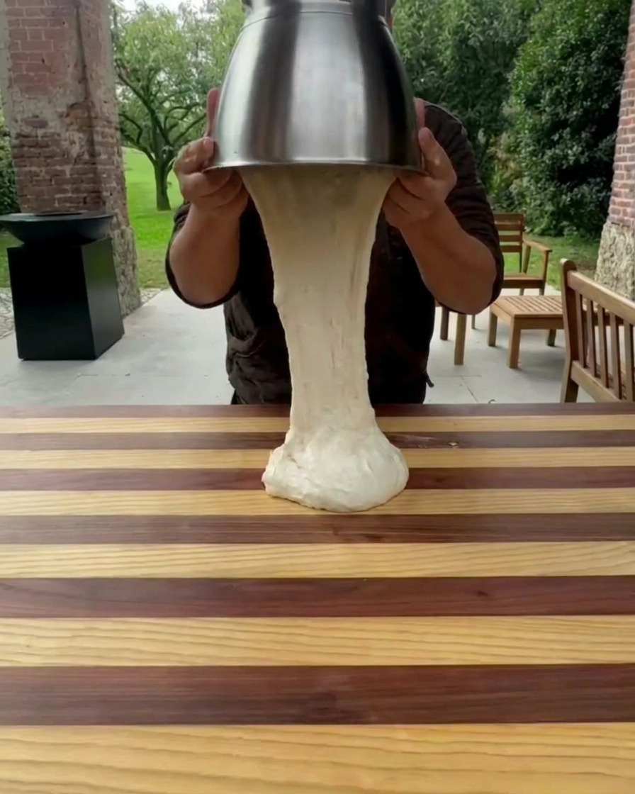 Dough being lifted out of a stainless steel mixing bowl onto a wooden countertop.