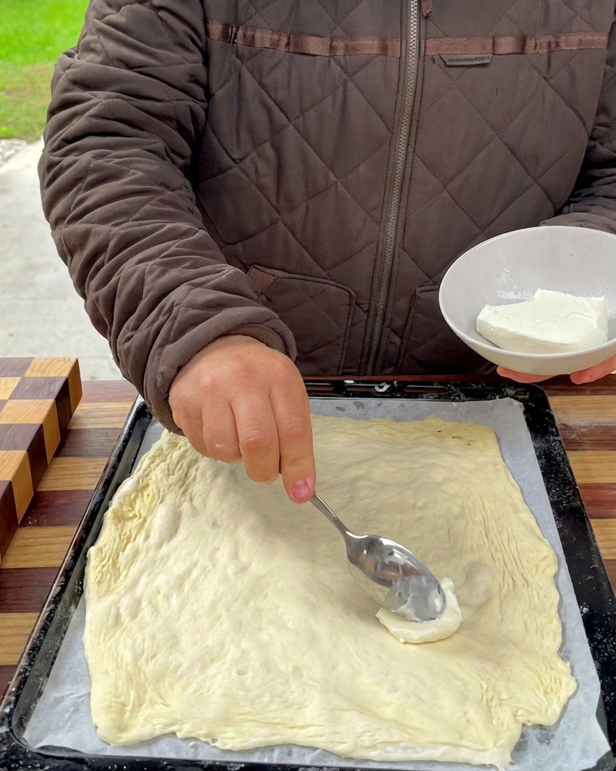 Creamy cheese base being spread across the surface of the pizza dough with a spoon.