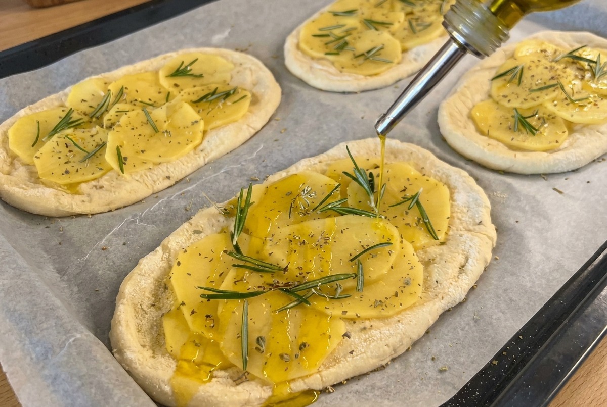 Olive oil being drizzled from a squeeze bottle onto potato-topped doughs, with salt being added from a grinder.