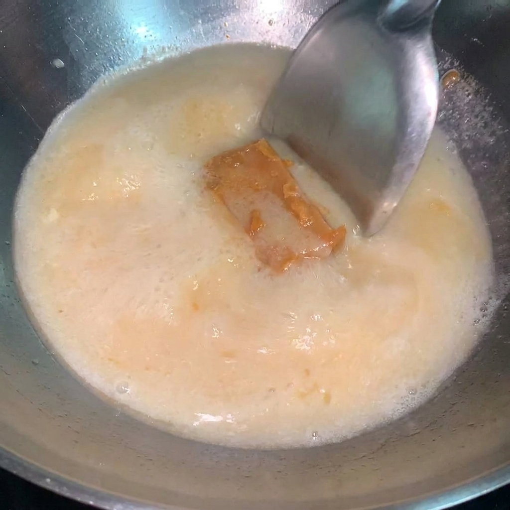 A spoonful of concentrated miso sauce being stirred into a simmering pot of ramen broth.