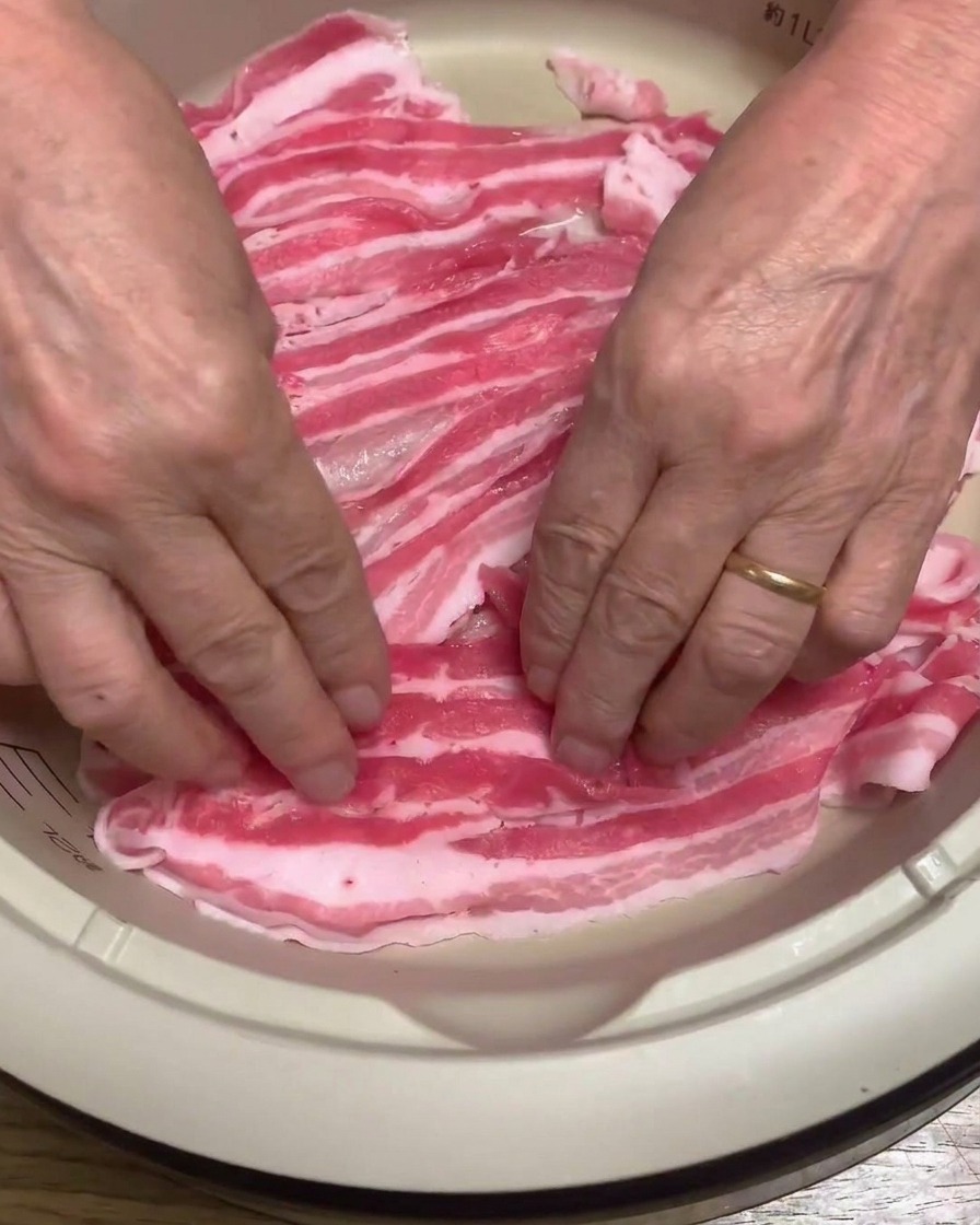 A close-up of raw pork belly slices being neatly arranged in the base of an electric pot.