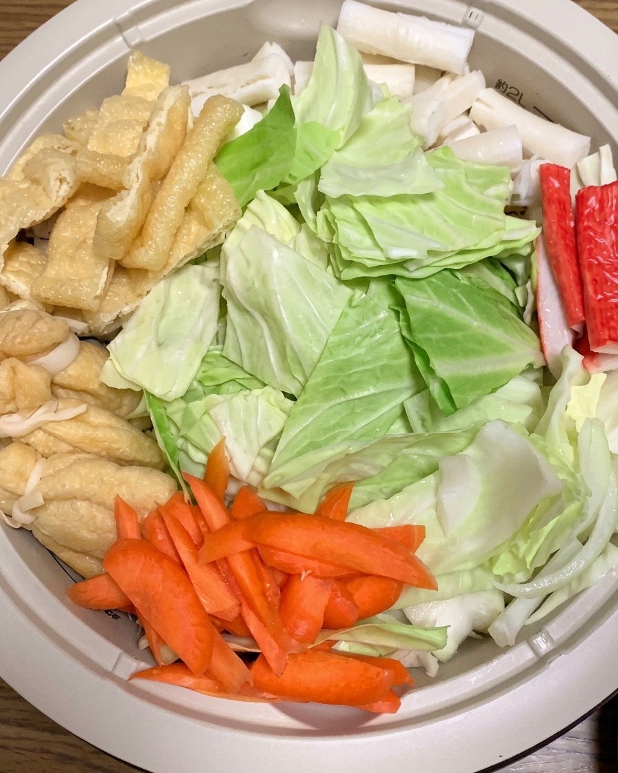 Hot water being poured into a pot filled with raw ramen ingredients including cabbage, carrots, and fried tofu.