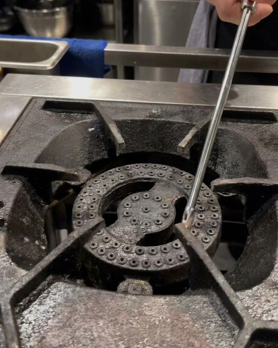 A hand using a long lighter to ignite a professional gas stove burner with a small pot sitting empty on the grate.