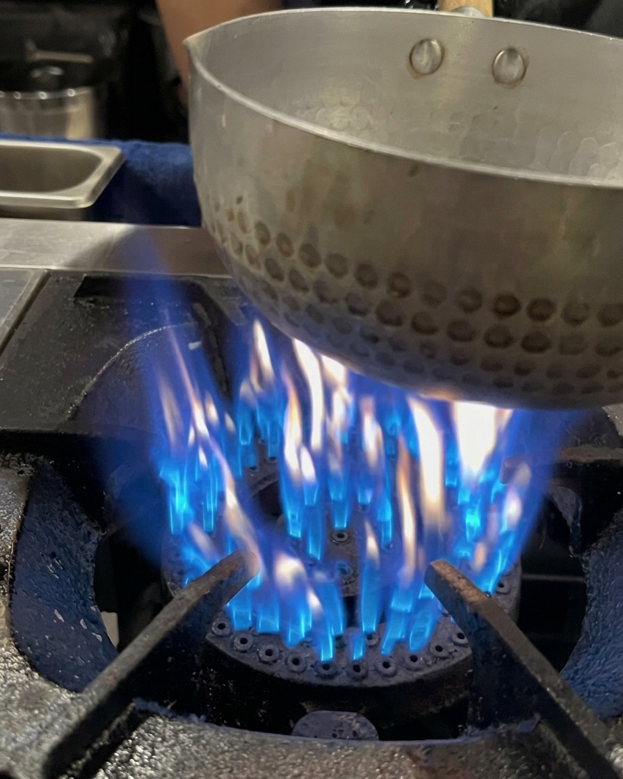 Pork bone broth being poured into a small pot sitting over an active blue gas flame.