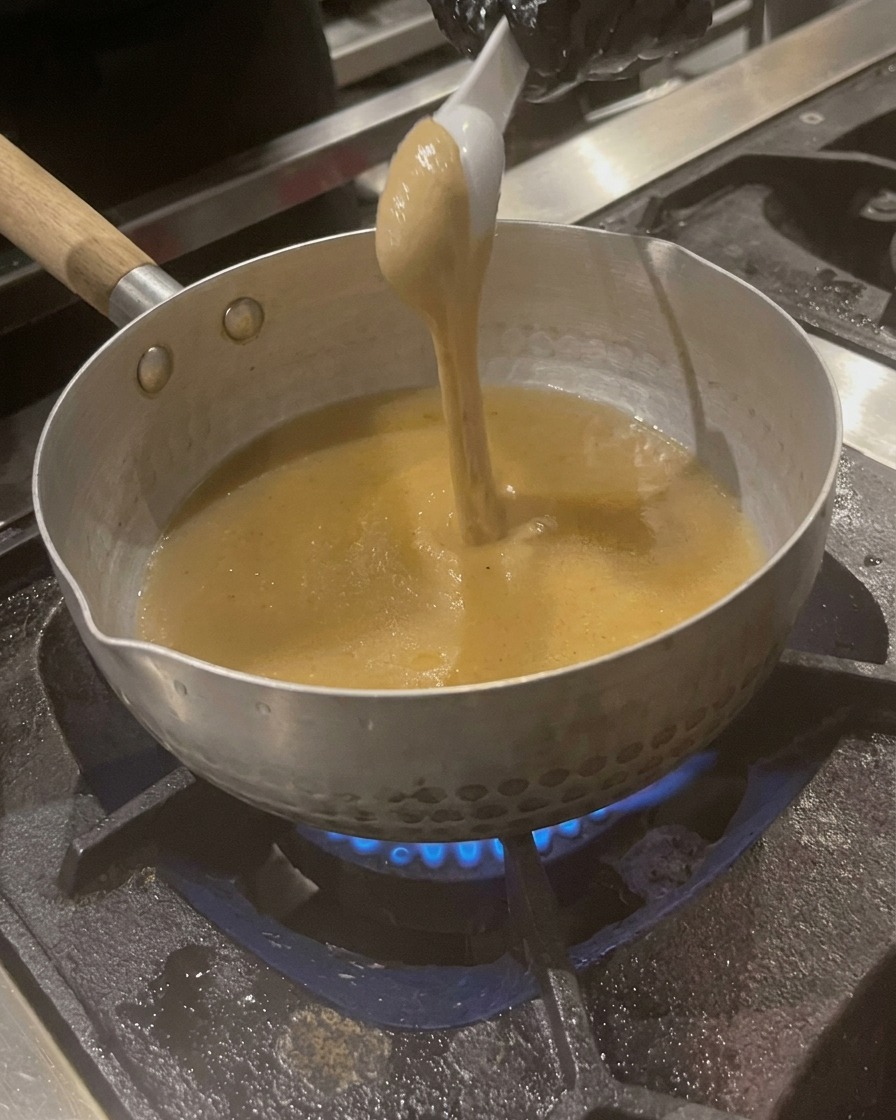 A spoonful of thick, tan-colored sesame paste being swirled into the simmering broth in the pot.