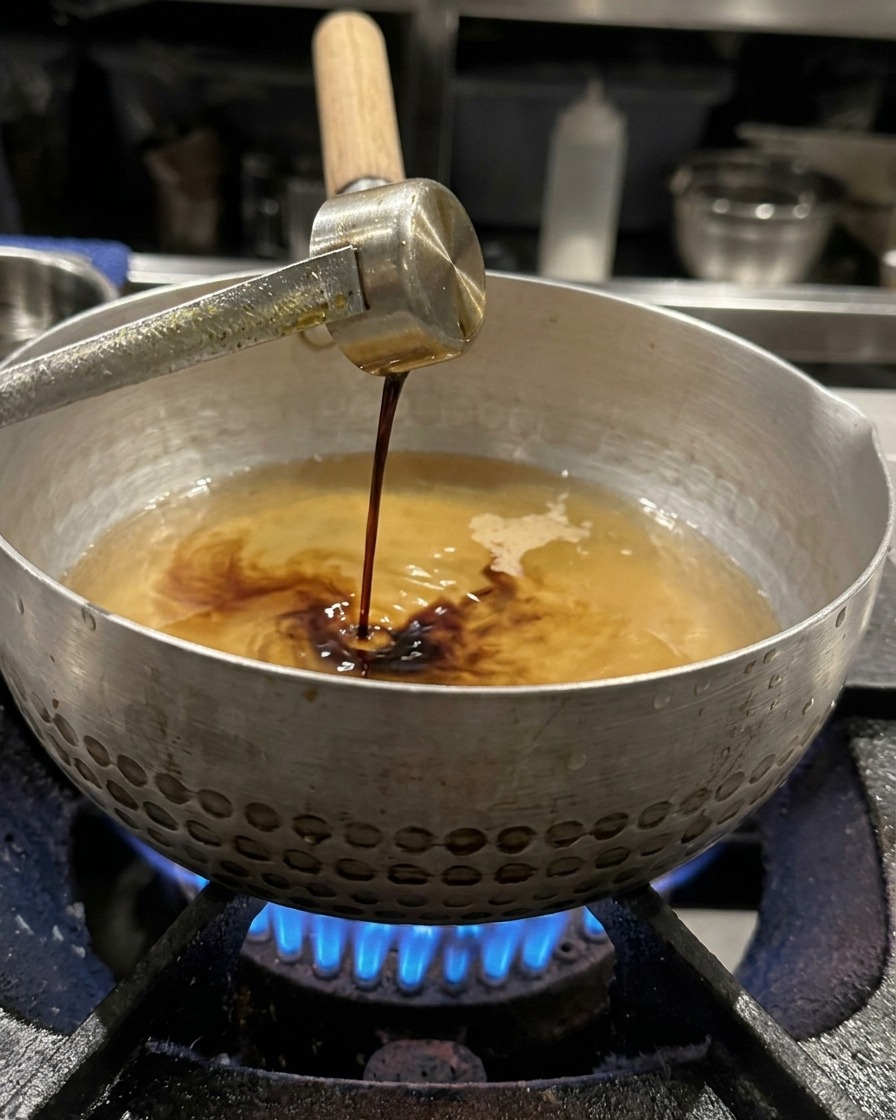 Soy sauce being drizzled from a metal ladle into the simmering broth, ready to be whisked.